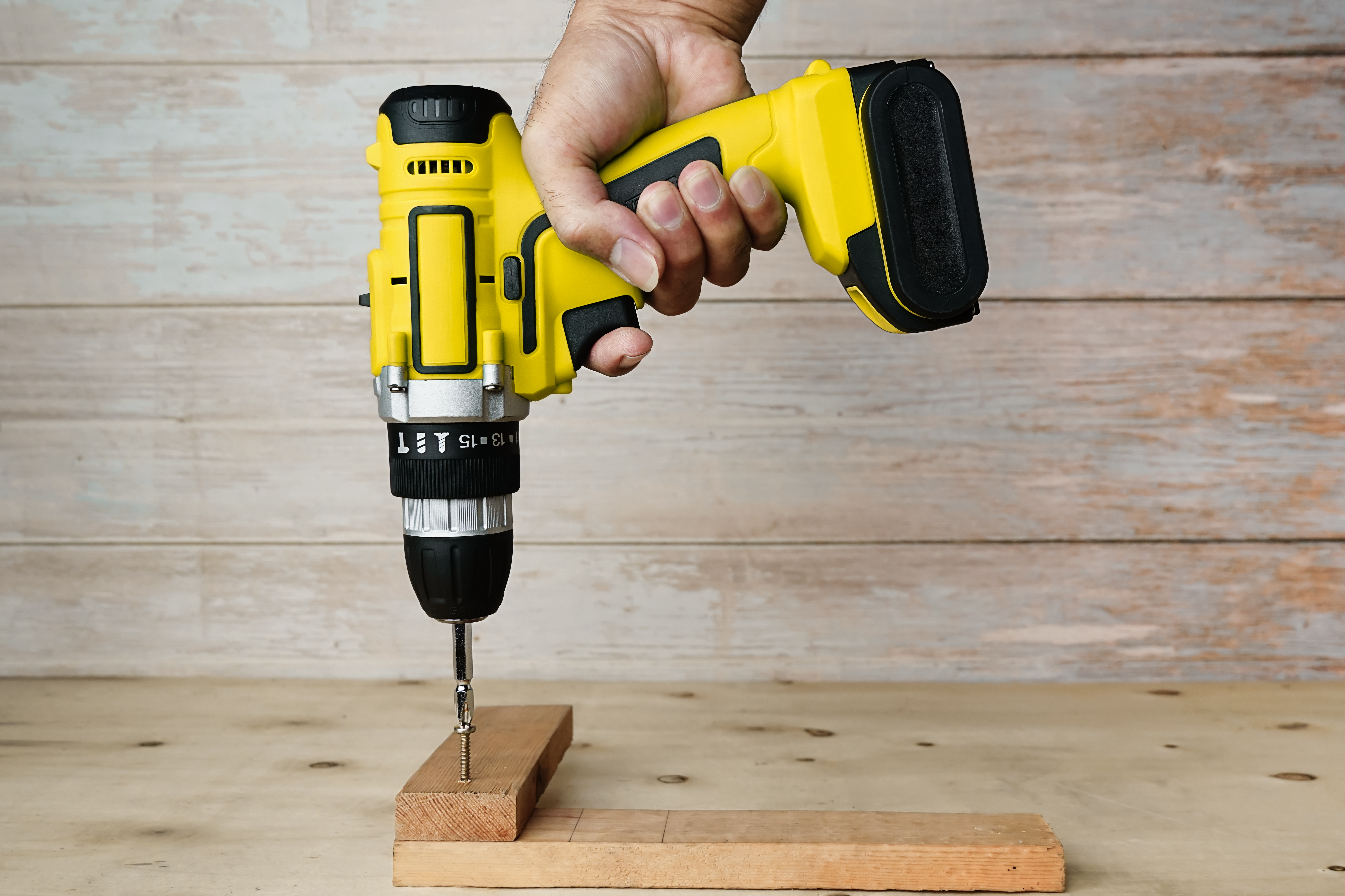 A close-up photograph of a carpenter's hand holding a cordless drill or electric screwdriver, screwing into a piece of wood