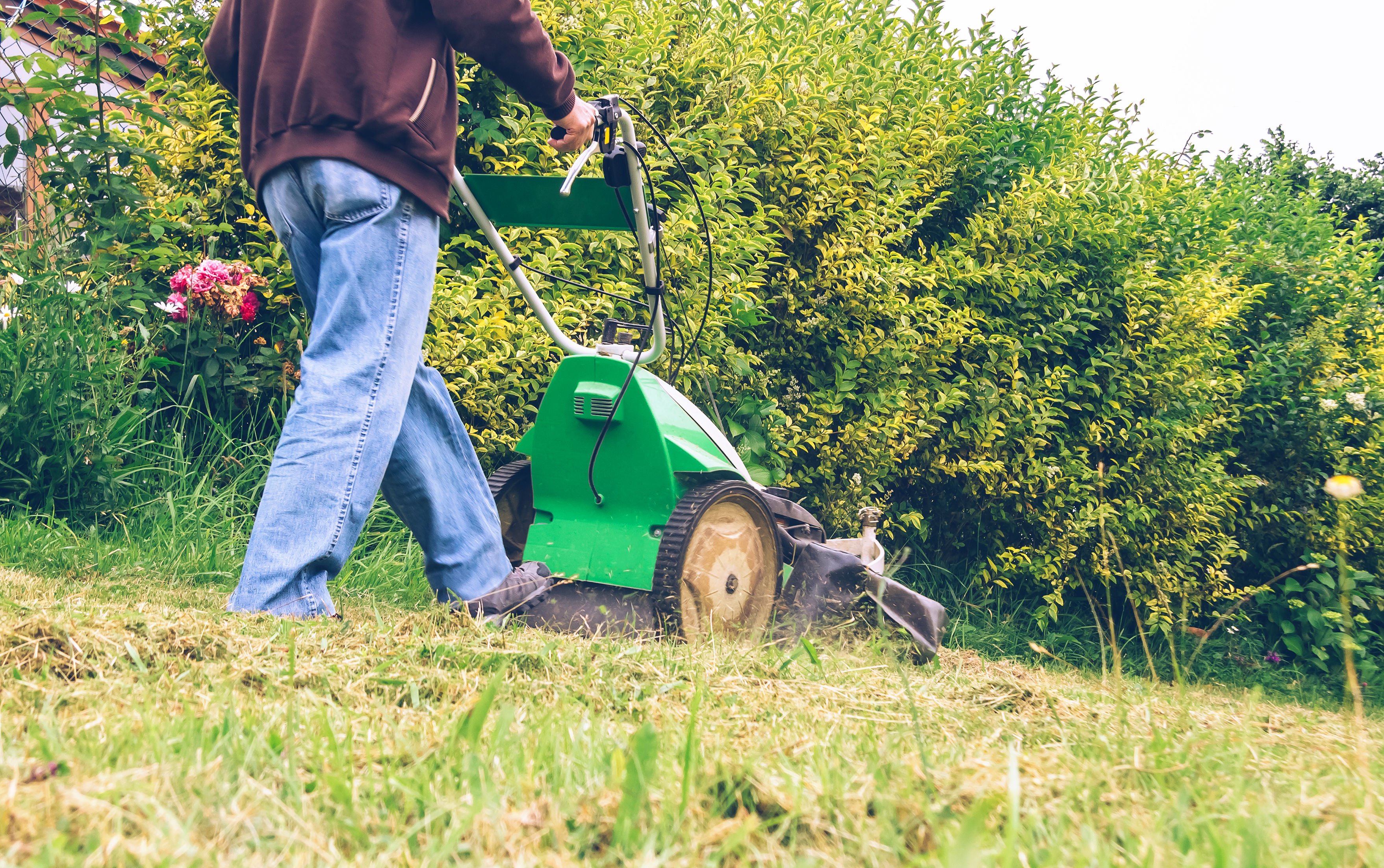 A close-up photograph of an elderly man wearing a cap, operating a lawnmower machine in a field or a large lawn, possibly in a rural or suburban setting
